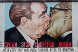 A photograph of Dmitri Vrubel’s mural, entitled ‘My God, Help Me to Survive this Deadly Love’. Framed by the title in both Cyrillic & German (painted in red and black block capitals) is a realist painting of Leonid Brezhnev and Erich Honecker kissing in front of a pale blue background.