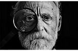 A monochrome image of an elderly man with tears welling in his eyes, looking through a magnifying glass with his right eye. The backdrop is black.