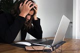 Woman in black jacket sits in front of an open laptop with her hands on her head, looking frustrated