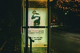 Photograph of a bus shelter in a leafy suburb at night with a light in its ceiling. A poster on a side wall of the shelter has the image of a young man being hugged by an invisible person. The poster’s caption reads “Over 170,000 people disappear every year”.