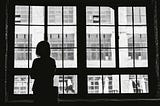 A silhouette of a person standing indoors, looking out through a large, multi-paned window. The window provides a view of the outside, where a brick building with numerous windows is visible. The scene is black and white, giving it a contemplative or nostalgic mood.