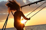 A woman swinging on a swing with the sun setting beyond the ocean’s horizon