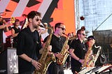 Coloured image showing several jazz musicians on stage, all wearing black shades and black clothing, and all playing saxophones and trumpets.