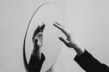 A black and white photo of a hand reaching towards and reflected in a mirror