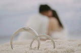 Two wedding rings in focus with a couple kissing in the background