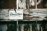 A worn, white picket fence with a white card nailed to it, reading: Resolutions. Photo by Tim Mossholder on Unsplash.