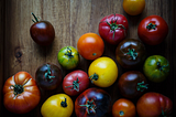 A rainbow of various sized tomatoes with most of them with green leaves and stems attached, placed on a wooden board.