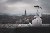 A small figure of a person made with snow, twigs and a leaf, sitting an a ledge high up, with a blurred view of a city in the distant background.
