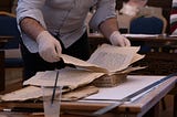 A conservator, wearing latex gloves, sorts the loose leaves of a manuscript book. Nearby are two metal rulers and a half-filled glass that contains a gray paste and an artist’s paintbursh.