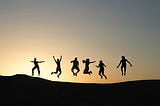 A coloured image showing six figures silhouetted on a hill against a rising or setting sun. The figures are jumping for joy.