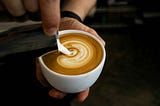 A cafe latte in someone’s left hand while the right hand pours cream.