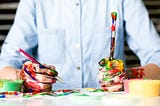 Person in button-down shirt sits at a table. The camera’s focus is on two hands covered in many colors of paint. One hand holds a mechanical pencil, the other a paintbrush. A neurodivergent woman’s life is messy and creative.