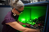 A researcher wearing safety glasses reaches into a box of circuitry and other equipment, which emits a green glow.
