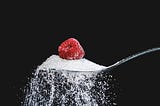 Close-up of an over-flowing silver teaspoon of sugar with a red raspberry on top, against a black background.