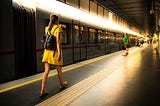 A train on the platform. A passenger in a yellow dress walks along the platform. In the end, more people are waiting.