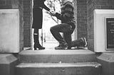 In grayscale a man kneels before a woman to propose at the top of a stairway between two brick walls.