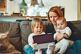 A mother sits on the couch holding her baby. Her toddler sits next to her. They are watching a tablet. Mother is pondering her next Medium article.