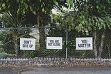 Fenceline backed by tropical trees with supportive signs on the fence that say “Don’t Give Up, “You Are Not Alone,” and “You Matter.”