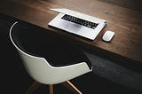 An extremely neat workplace — white chair at a dark-wood desk. A white laptop and a white mouse neatly laid out on the desk.