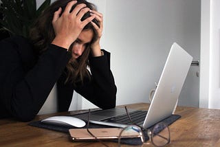 Woman in black jacket sits in front of an open laptop with her hands on her head, looking frustrated