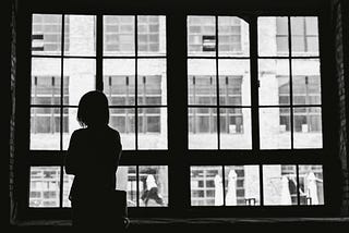 A silhouette of a person standing indoors, looking out through a large, multi-paned window. The window provides a view of the outside, where a brick building with numerous windows is visible. The scene is black and white, giving it a contemplative or nostalgic mood.