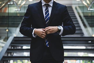 Image of businessman in full suit and tie as seen from the neck down, standing in front of open staircase and adjusting suit jacket.