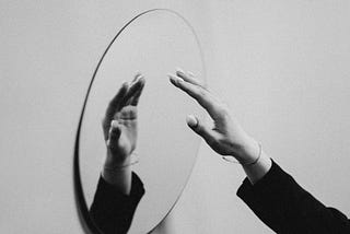 A black and white photo of a hand reaching towards and reflected in a mirror