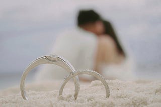 Two wedding rings in focus with a couple kissing in the background
