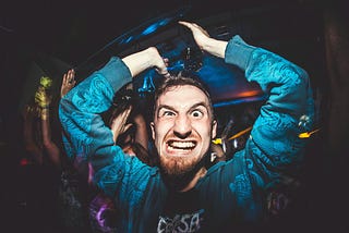 A young, bearded, man at a party raises his arms above his head as he stares at the viewer with a crazy-looking expression!