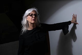 older woman with long white hair leans an arm against a wall, looking wary