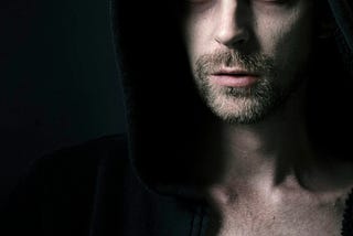 A the face of a man with very pale skin, red eyes and scratches on his head. His head is covered with a dark hoody.