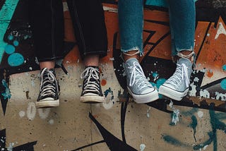 Two pairs of sneaker-clad feet belonging to people hanging out sitting on a wall with grafitti on it.
