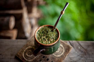 A ceramic cup of mate sits on a wooden table. It is full of a drink with a lot of green herbs floating on the top, and a spoon sticking out of it.