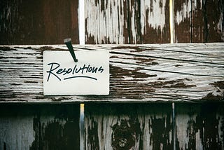 A worn, white picket fence with a white card nailed to it, reading: Resolutions. Photo by Tim Mossholder on Unsplash.