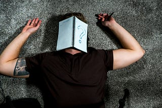 This is a photo of a man lying on the floor with his face covered by a notebook on which a question mark has been drawn.