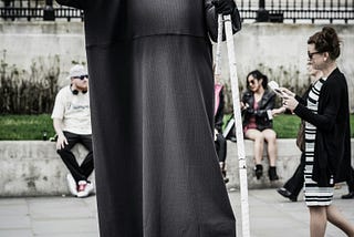 A coloured image showing a street performer dressed as the Grim Reaper. He is holding a scythe in his left hand. People sit on a low wall behind. A woman walks past him. No one is paying him any attention.