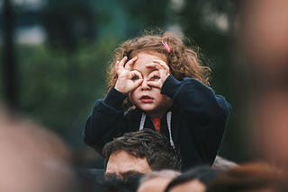 Little girl on her daddy’s shoulders with her fingers cupped around her eyes, mimicking eyeglasses.