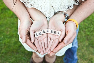 A picture of a parent holding a childs hands while the child holds the scrabble letters spelling “forever’