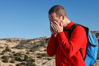A man in a red shirt wears a backpack and holds his hands to his face