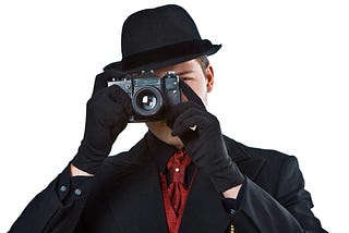 Man in suit, hat and gloves holding a camera in front of his face to take a picture.