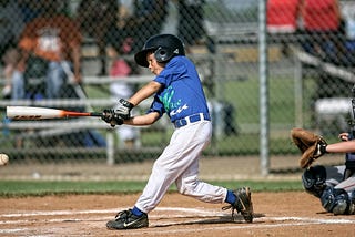 A young boy swinging his bat while at home plate getting pitches from the opposite team’s pitcher. The catcher is behind the young man. Swinging at baseballs allows the player to have three strikes before they are called out.