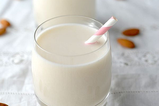 A photograph of two glasses of almond milk with pink and white straws. They are placed on a white tablecloth with lace inserts and almonds without shells are placed randomly across the tablecloth. The background is blurred.