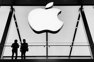 A large, illuminated Apple logo prominently displayed in a store. Below the logo, two individuals are standing at a balcony, likely within the Apple Store. The black and white tones enhance the modern and sleek aesthetic typical of Apple’s store design. This setting, with its sharp contrasts and iconic logo, effectively highlights the branding and architectural elements that Apple is known for.