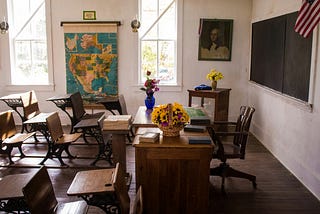 Country classroom with student desk, teacher’s desk, and map on the wall.