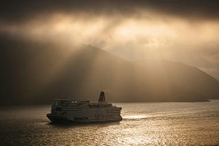 a cruise ship or ferry boat sailing on the ocean in the mist- you can see the sun peeking through the clouds and mountains in the background