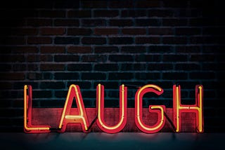 Coloured image in which a neon sign spells out the word ‘laugh’ in bright letters against a dark brick wall.