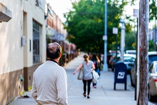 A coloured image showing a middle-aged man in the foreground, back to the camera, watching as a woman walks away from him down the street. She’s carrying two bags.