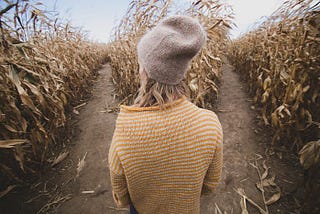 Photo taken from the back of a young woman standing between the rows of dried cornstalks, her shoulders slumped, staring in sad contemplation.