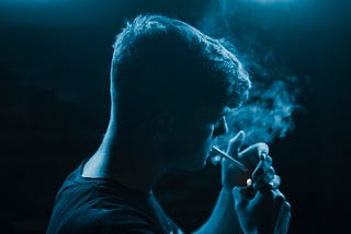 A male person in blue light lighting a cigarette.