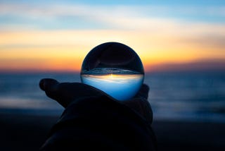 A dark hand holding a glass sphere in front of a beach. The image in the sphere is darker than the background.